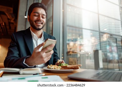 Smiling Indian businessman wearing suit is using phone during lunch time in cafe. Blurred background - Powered by Shutterstock