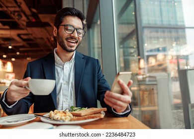 Smiling Indian businessman wearing suit is using phone during lunch time in cafe. Blurred background - Powered by Shutterstock