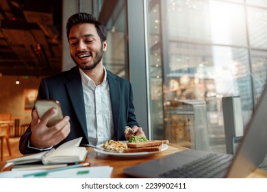 Smiling Indian businessman wearing suit is using phone during lunch time in cafe. Blurred background - Powered by Shutterstock