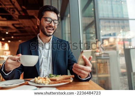 Smiling Indian businessman wearing suit is using phone during lunch time in cafe. Blurred background