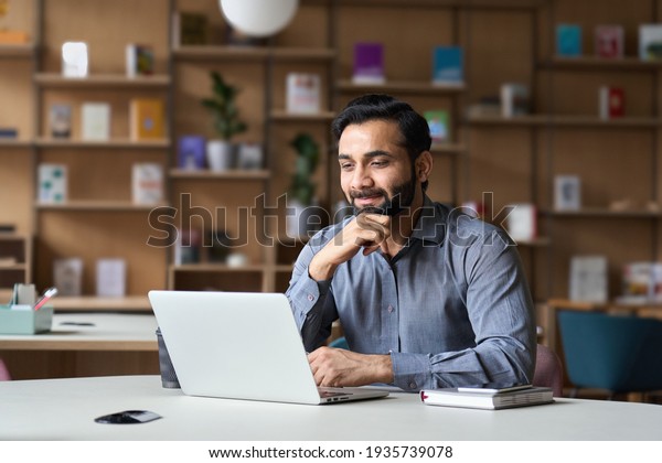 Smiling indian businessman working on laptop in\
modern office lobby space. Young indian student using computer\
remote studying, watching online webinar, zoom virtual training on\
video call meeting.