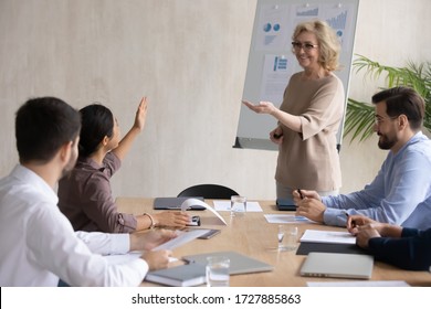 Smiling Indian Businesswoman Raising Hand Asking Mature Mentor In Boardroom At Company Meeting. Diverse Happy Woman Employee In Conversation Asks Older Lead Question About Business Sit At Table.