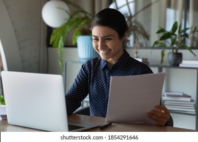 Smiling Indian businesswoman using laptop, checking financial documents, correspondence, happy employee holding paper sheet, looking at computer screen, satisfied by good news, great project results - Shutterstock ID 1854291646