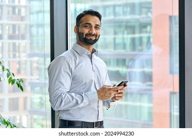 Smiling indian ceo businessman holding using cell phone mobile apps standing in office near panoramic window looking at camera. Digital tech apps and solutions for business corporate development.