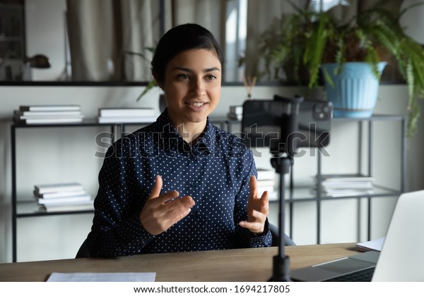 Smiling indian ethnic girl sitting in front of\
smartphone on stabilizer, recording self-presentation video or\
sharing professional skills. Happy young smart businesswoman\
filming educational\
lecture.