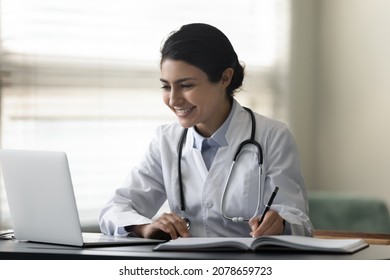 Smiling Indian female doctor physician in uniform with stethoscope using laptop, taking notes, writing in medical journal, professional therapist practitioner filling documents or watching webinar