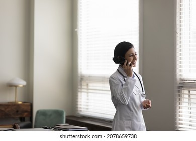 Smiling Indian female doctor physician in white uniform with stethoscope talking on cellphone standing in medical office, confident therapist general practitioner consulting patient by phone call
