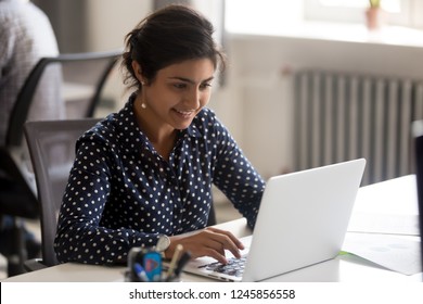 Smiling Indian female employee using laptop at workplace, looking at screen, focused businesswoman preparing economic report, working online project, cheerful intern doing computer work, typing