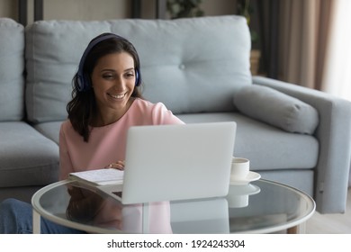 Smiling Indian Female Freelancer Wearing Headset Working In Comfort Living Room Look At Pc Screen Talking By Video Call. Young Arabian Woman Study From Home On Distance Take Part In Conference Webinar