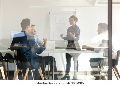 Smiling indian female presenter speaker give flip chart presentation in boardroom behind glass door, happy hindu business coach training diverse team talking to workers group at office workshop