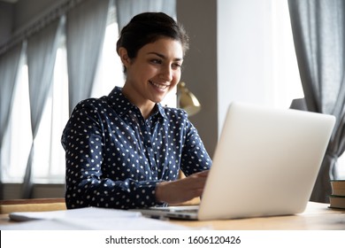 Smiling indian girl student professional employee typing on laptop sit at home office table, happy hindu woman studying e learning online software using technology app for work education concept