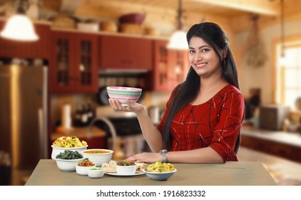 Smiling Indian Girl Or Woman Holding A Empty Bowl Sitting In Relax Mood Beside Group Of Traditional Indian Dishes And Looking At The Camera. Female Model Potrait