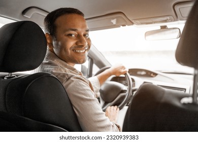 Smiling indian guy driver sitting inside brand new car, looking at back seat, copy space. Happy arabic millennial man enjoying driving comfortable automobile, going on summer vacation