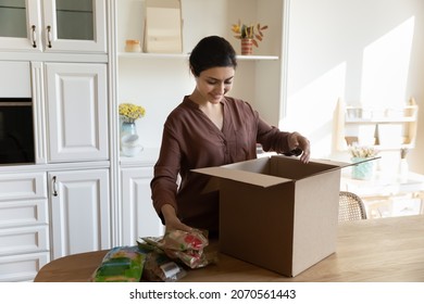 Smiling Indian housewife woman standing in modern kitchen takes out of box ordered products received order from courier. Express comfort food delivery services, online remote grocery shopping concept