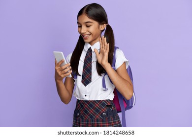 Smiling indian latin kid girl wearing uniform holding phone, waving hand, video calling friend, family or school teacher during virtual meeting remote distance learning isolated on lilac background.