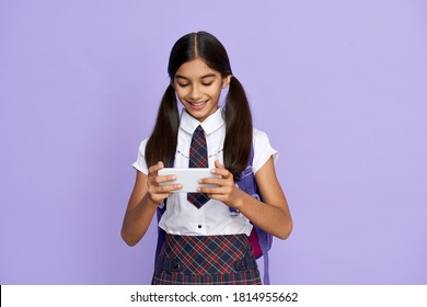 Smiling Indian Latin Preteen Kid Girl Wears School Uniform Holds Backpack And Phone Watching Mobile Streaming Ad, Remote Learning Online Class, Video Calling On Cellphone Isolated On Lilac Background.