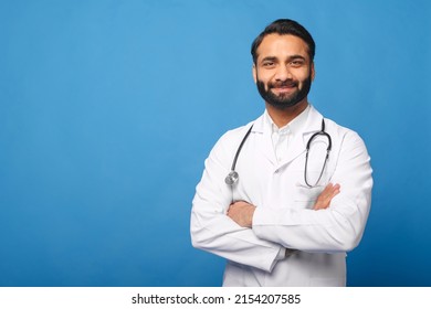 Smiling Indian male doctor pediatric, physical, therapist wearing white medical gown with stethoscope on shoulders stands with arms crossed isolated on blue, copy space. Healthcare and medicine