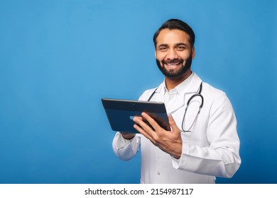 Smiling Indian Male Doctor Pediatric, Physical, Therapist Wearing White Gown With Stethoscope On Shoulders Using Digital Tablet For Online Communication With Patients Isolated On Blue, Telemedicine