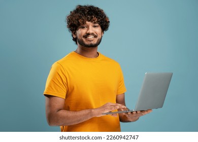 Smiling Indian man holding laptop computer working online looking at camera. Happy confident university student studying isolated on yellow background, online education. Portrait of happy programmer  - Powered by Shutterstock