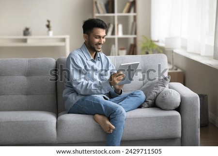 Smiling Indian man spend pastime on internet sit on couch with modern digital tablet, using new program, enjoy hobby, do electronic shopping via marketplace apps or web sites, e-dating services user