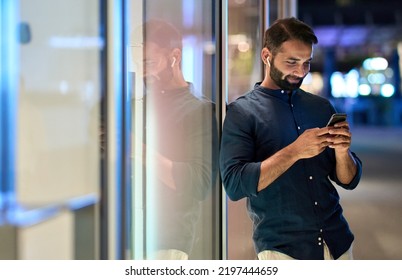 Smiling Indian Man Wearing Earbud Holding Mobile Phone Listening Music At Night Outdoors, Using Smartphone Having Chat Call In App Tech On Cellphone, Watching Streaming Tv Videos Online.