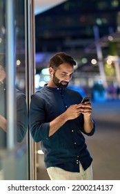 Smiling Indian Man Wearing Earbud Holding Mobile Phone Listening Music At Night Outdoors, Using Smartphone Having Chat Call In App Tech On Cellphone, Watching Streaming Tv Videos Online, Vertical.