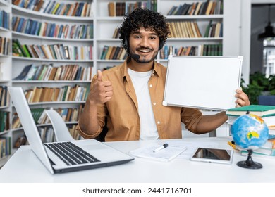 Smiling indian man with wireless headset holding blank whiteboard and giving thumbs up in front of modern laptop with various items on desk. Background of bookshelves and home office.