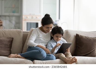 Smiling Indian mother with son using tablet at home, relaxing sitting on cozy couch together, loving young mom and 5s boy child looking at gadget screen, watching video or cartoons, playing games - Powered by Shutterstock