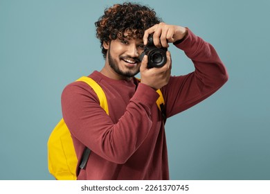 Smiling Indian photographer holding photo camera taking pictures isolated on blue background.  Attractive asian tourist with backpack looking at camera. Travel concept