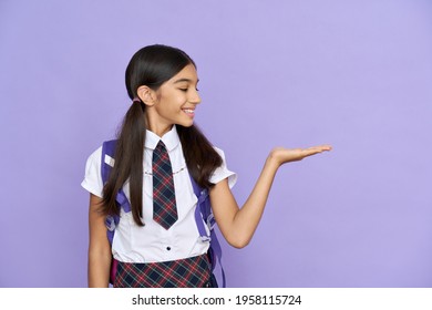 Smiling indian schoolgirl, preteen kid smiling looking at copy space holding hand. Advertising latin junior student standing isolated on violet lilac background. Education, school concept.