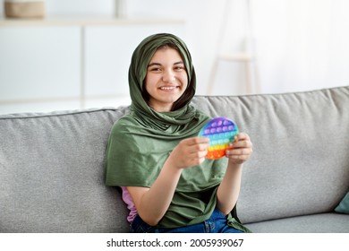 Smiling Indian Teen Girl In Hijab Sitting On Comfy Couch, Playing With Trendy POP IT Toy At Home. Muslim Adolescent In Traditional Headscarf Enjoying Antistress Plaything, Popping Bubbles Indoors