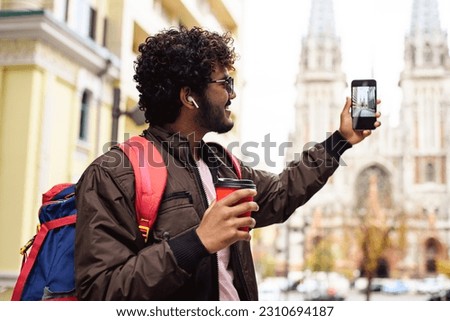 Smiling indian traveler with coffee to go taking photo on smartphone on urban street	
