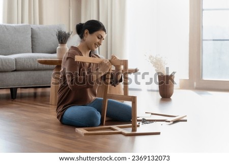 Smiling Indian woman assembling new wooden furniture, sitting on floor in living room at home, happy young female renter customer putting wooden shelf or chair pieces together, renovation concept