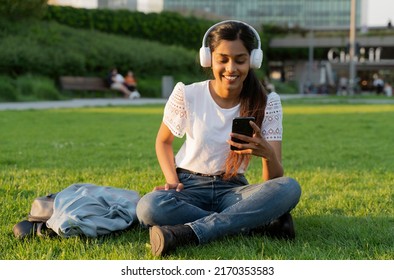 Smiling Indian woman holding mobile phone listening music in wireless headphones sitting on grass. Happy asian hipster using mobile app shopping online, reading text message in park