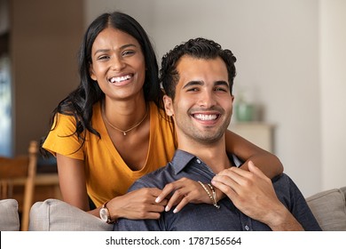 Smiling Indian Woman Hugging Her Husband On The Couch From Behind At Home. Loving Middle Eastern Couple Looking At Camera With Big Grin. Portrait Of Laughing Girl Embracing Handsome Latin Man On Sofa.