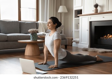 Smiling Indian woman practicing yoga online using laptop, working out at home, happy young female in grey sportswear doing upward facing dog pose, stretching in Urdhva mukha shvanasana exercise - Powered by Shutterstock