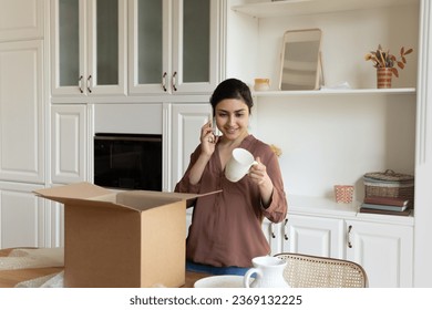 Smiling Indian woman talking on phone, chatting with friend, unpacking box with fragile online store order, holding porcelain cup, happy young female unboxing parcel with belongings in new apartment