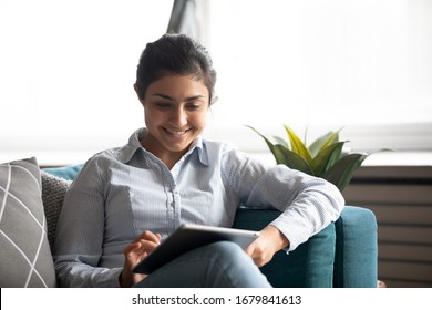 Smiling Indian Woman Using Computer Tablet, Sitting On Cozy Couch In Living Room, Happy Girl Looking At Screen, Watching Funny Video, Chatting In Social Network Or Shopping Online, Playing Game