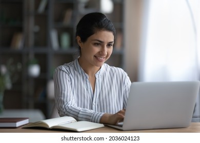 Smiling Indian Woman Using Laptop, Studying Online At Home, Sitting At Desk With Books And Computer, Working On Research Project Or Homework, Positive Businesswoman Writing Report Or Email