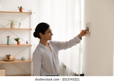 Smiling Indian woman using modern smart home system, controller on wall, positive attractive young female switching temperature on thermostat or activating security alarm in apartment