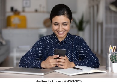 Smiling Indian Woman Using Smartphone, Sitting At Home Office Desk, Happy Young Female Businesswoman Freelancer Or Student Typing On Phone Screen, Chatting Online In Social Network During Break