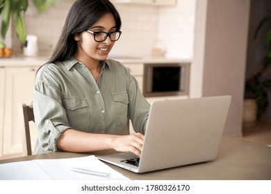 Smiling indian young adult woman wearing glasses typing on laptop computer working at home office sitting at table. Happy female professional freelancer student studying online using notebook pc.