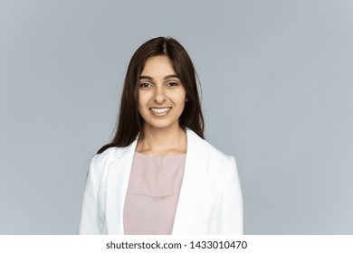 Smiling indian young businesswoman wear white suit looking at camera isolated on grey studio background, happy attractive confident hindu business lady professional head shot close up portrait