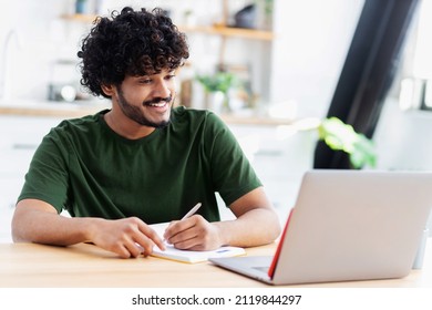 Lächelt indische junge Mann mit Laptop zum Anschauen von Webinar, studieren online und nimmt Notizen in einem Notizbuch, männliche Schüler studiert online, E-Learning-Konzept