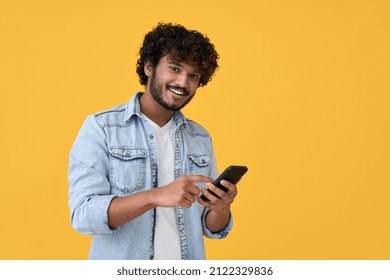 Smiling indian young man using cell phone isolated on yellow background. Happy guy holding smartphone ordering online, making mobile banking payment, advertising application on cellphone.