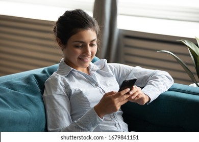 Smiling Indian Young Woman Using Smartphone, Sitting On Cozy Couch At Home, Satisfied Happy Girl Holding Phone, Looking At Screen, Chatting In Social Network, Surfing Internet, Spending Leisure Time