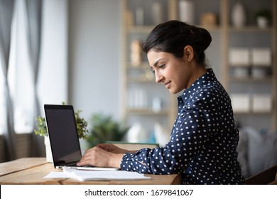 Smiling Indian young woman working on laptop, reading documents, successful female freelancer writing project report, using computer, focused girl student preparing to exam, sitting at desk at home - Shutterstock ID 1679841067