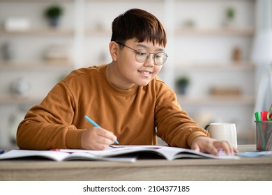 Smiling intelligent asian schooler chubby teen boy doing homework, sitting at table and writing at notebook, enjoying educational process, copy space, home interior. Kids and school concept