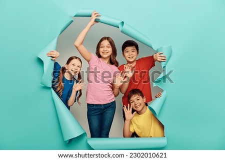 Smiling interracial kids in casual clothes waving hands at camera during international children day celebration behind hole in blue paper background