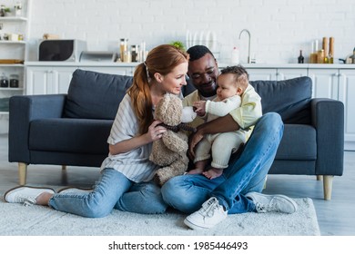 smiling interracial parents sitting on floor with infant daughter and teddy bear - Powered by Shutterstock